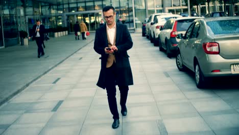 A-businessman-walks-with-a-mobile-phone-at-a-business-center,-close-up.