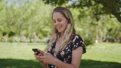 Cheerful-attractive-blonde-young-woman-standing-in-the-park-with-earphones-in-her-ears-using-mobile-phone-smiling-and-happy