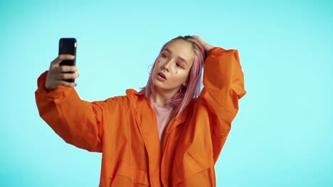 Girl-with-pink-hair-and-extraordinary-appearance-make-selfie-on-blue-background.-Using-modern-technology---smartphone,-social-networks