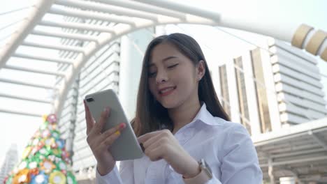 Joven-mujer-asiática-sonriente-de-negocios-usando-teléfono-inteligente-en-el-centro-de-la-ciudad-moderna-en-la-noche-de-Bangkok-Tailandia.-Comunicación-Concept-Technology-por-teléfono-móvil