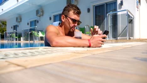 Mann-mit-Telefon-am-Pool-und-erfrischenden-kalten-Cocktail.-Guy-trinken-Getränke-Surfen-Internet-Seite-oder-kommunikation-mit-jemandem.-Junger-Mann-genießt-Erholung-auf-Resort-während-der-Sommerferien.-Niedrige-Ansicht