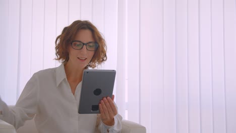 Closeup-portrait-of-adult-redhead-caucasian-businesswoman-in-glasses-having-a-video-call-on-the-tablet-waving-hi-sitting-in-the-armchair-in-the-white-office