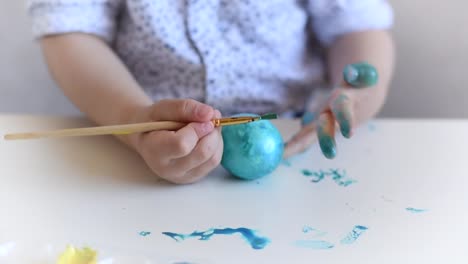 The-hands-of-a-litlle-child-with-a-brush-paint-an-Easter-egg-in-blue-color-on-the-table.