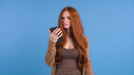 Rothaarige-Mädchen-in-braunem-T-shirt-und-Strickjacke.-Sie-benutzt-das-Smartphone,-schaut-genervt-und-fragt-wtf,-während-sie-vor-blauem-Hintergrund-posiert.-Nahaufnahme