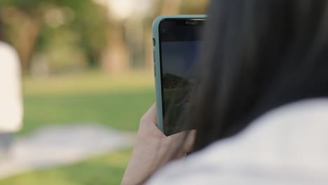 Schöne-Teenager-Mädchen-mit-Smartphone-machen-ein-Foto-mit-Freunden,-während-sie-auf-dem-Boden-Gras-im-Freien-in-einem-öffentlichen-Park-auf-dem-schönen-Sonnenuntergang-sitzen.