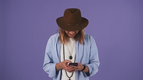 Long-haired-fellow-in-casual-clothes,-glasses,-hat.-Smiling,-using-smartphone,-looking-surprised-and-saying-wow.-Posing-on-purple-background.-Close-up