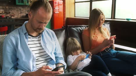 Papá,-mamá-y-su-hijo-en-ropa-casual-están-usando-teléfonos-móviles-mientras-están-sentados-en-el-sofá-en-el-apartamento-estudio-con-interior-elegante.-Cámara-lenta