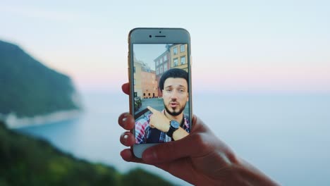 Close-up-of-hand-holding-smartphone-during-video-call-with-man-in-front-of-the-sea