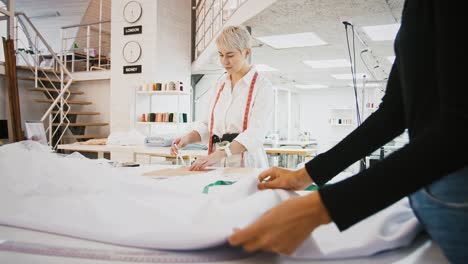 Woman-tailor-of-sewing-workshop-drawing-pen-around-pattern-standing-at-table,-seamstress-spreading-white-fabric