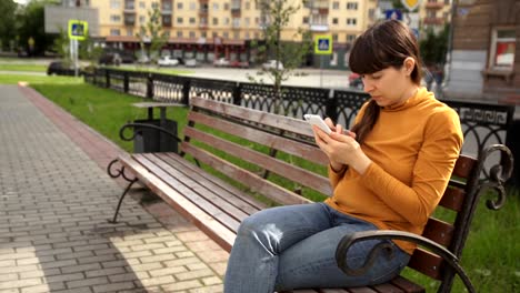 Young-woman-using-telephone-sitting-on-the-bench-near-the-road