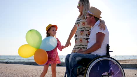 disabled-person-in-wheelchair-with-family,-motherhood,-little-girl-listens-mother-and-father-on-beach-in-summer-time,-happy-mom-and-daughter-stand-near-dad