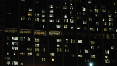 Office-building-in-the-dark.-The-windows-are-lit,-people-silhouettes-are-visible.-Tilt-shot