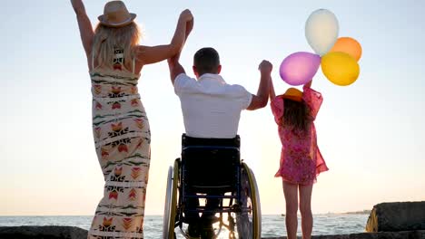 Happy-rest,-invalid-with-wife-and-daughter-raise-their-hands-up-with-colorful-air-balloons-near-sea,-family-with-child