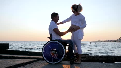 disabled-man-caresses-woman-with-big-tummy-on-embankment,-disabled-man-in-wheelchair-listens-belly-of-pregnant-woman