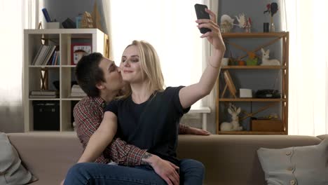 Zwei-junge,-schöne-Mädchen-auf-der-Couch-sitzen,-küssen-und-machen-Selfies,-Lesben,-LGBT,-blond,-brünett-60fps