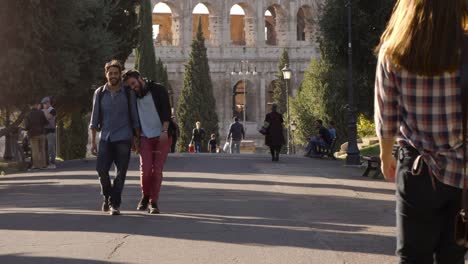 Young-happy-gay-couple-tourists-walk-in-park-road-with-trees-colosseum-in-background-in-rome-at-sunset-holding-hands-lovely-slow-motion-colle-oppio