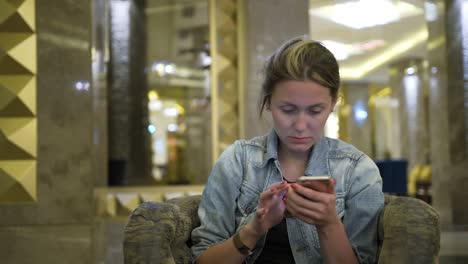 mujer-joven-caucásica-en-una-chaqueta-de-jeans-bebe-jugo-de-un-vaso-y-utiliza-un-teléfono-inteligente,-escribe-los-mensajes-en-la-red-social.-Se-sienta-en-la-mesa-en-el-restaurante-del-hotel.-Concepto-de-sano-alimento-fresco-en-los-negocios.-Cierre-para-