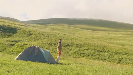 Tourist-man-came-out-of-camping-tent-on-green-meadow-and-mountain-background