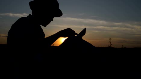 Silhouette-eines-jungen-verwendet-eine-Tablette-bei-Sonnenuntergang-im-Feld-liest-etwas-auf-dem-tablet