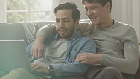 Sweet-Male-Queer-Couple-Spend-Time-at-Home.-They-are-Lying-Down-on-a-Sofa-and-Use-the-Laptop.-They-Browse-Online.-Partner's-Hand-is-Around-His-Lover.-They-Smile-and-Laugh.-Room-Has-Modern-Interior.