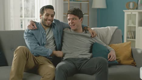 Slow-Motion-of-a-Male-Gay-Couple-Fulling-Around-on-a-Sofa-at-Home.-Boyfriend-Runs-and-Jumps-into-Hands-of-His-Partner.-They-Hug.-They-are-Happy-and-Laughing.-They-are-Casually-Dressed-and-Room-Has-Modern-Interior.