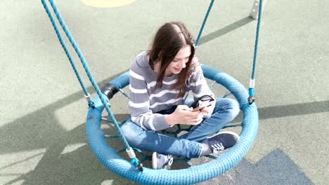 Beautiful-brunette-girl-is-swinging-on-empty-webbed-swing,-typing-a-message-on-mobile-phone.