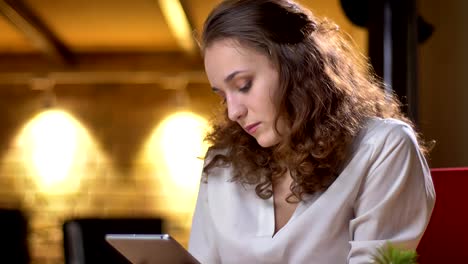 Portrait-of-young-curly-haired-businesswoman-attentively-watching-into-tablet-and-raising-her-eyes-to-camera-in-office.