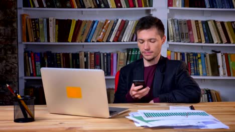 Attraktiver-männlicher-Student-sitzt-vor-dem-Laptop-und-stöbert-am-Telefon-in-der-Bibliothek-drinnen