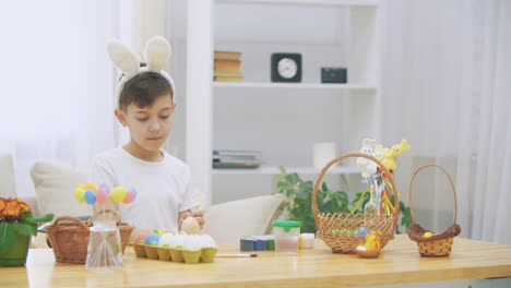 Creative-boy-is-colorizing-an-Easter-egg-with-a-help-of-paint-brush.,-wearing-bunny-ears,-then-he-takes-a-wooden-basket-and-shows-how-many-coloured-Easter-eggs-there-are,-and-points-with-his-finger-how-many-eggs-he-has-coloured.