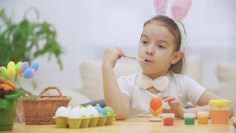 Little-playful-girl,-wearing-bunny-ears-on-her-head-chose-a-red-colour-to-paint-an-egg.-Girl-has-painted-an-heart-on-it.-Girl-is-cleaning-her-nose-and-is-putting-a-paint-brush-near-to-her-mouth-and-face.