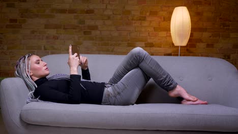 Closeup-shoot-of-young-attractive-caucasian-hipster-female-using-the-tablet-happily-smiling-lying-on-the-couch-indoors-in-a-cozy-apartment