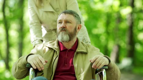 Zoom-in-of-female-caregiver-pushing-man-in-wheelchair-towards-camera-in-park.-Tired-senior-man-looking-at-camera