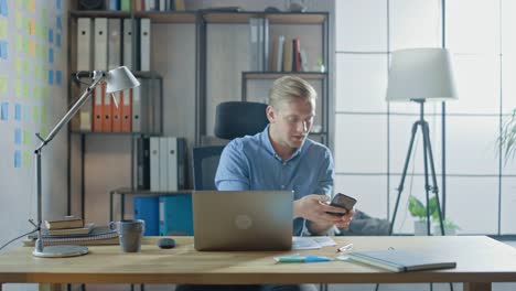 Entrepreneur-Sitting-at-His-Desk-Works-on-Desktop-Computer-in-the-Stylish-Office,-Checks-His-Smartphone,-Social-Media-App,-email-Business-Associates,-Messages