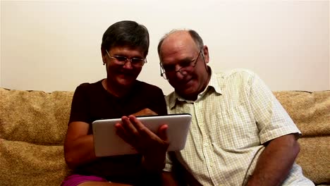 Una-pareja-de-ancianos-está-sentada-en-un-sofá-en-casa-y-viendo-fotos-en-una-tableta-PC,-sonriendo-y-hablando-activamente.