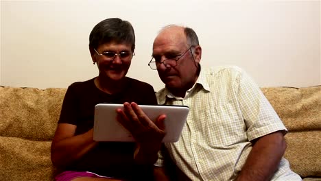 An-elderly-couple-is-sitting-on-a-sofa-at-home-and-watching-photos-on-a-tablet-pc,-smiling-and-talking-actively.