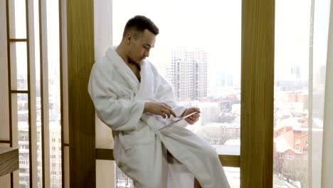 Man-dressed-in-white-bathrobe-sitting-on-windowsill,-using-tablet-computer.-4K