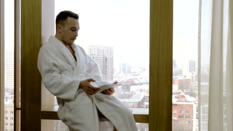 Man-dressed-in-white-bathrobe-sitting-on-windowsill,-using-tablet-computer.-4K