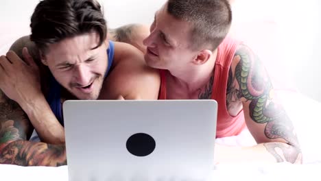 Gay-couple-in-bed-using-laptop-computer.-Kissing-in-cheek.