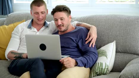 Pareja-gay-relajándose-en-el-sofá-usando-la-computadora-portátil.-Navegación.