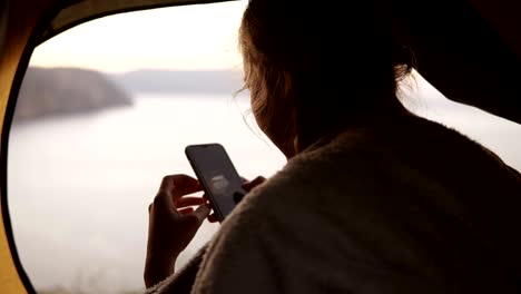 Camping-woman-is-sitting-in-tent-watching-taken-photos-of-nature-on-her-smart-phone.-Camping-girl-relaxing-on-vacation.-Sea-or-lake-from-the-top-of-a-hill-on-background