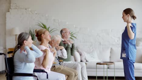 Side-view-tracking-shot-of-young-nurse-and-group-of-senior-patients-including-disabled-ones-doing-arm-rotation-exercise-together-in-nursing-home