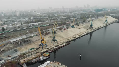 Aerial-view-of-cargo-port-cranes-near-a-pile-of-sand-with-ships,-scows-and-barges-transporting-river-sand.-Industrial-city-covered-with-smog-and-fog