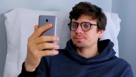 Sleepy-man-in-glasses-yawns-and-browsing-in-social-media-on-smartphone-sitting-in-armchair.