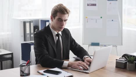 Geschäftsmann-SMS-geheime-Informationen-mit-Handy-während-der-Arbeit-auf-Büro-Laptop