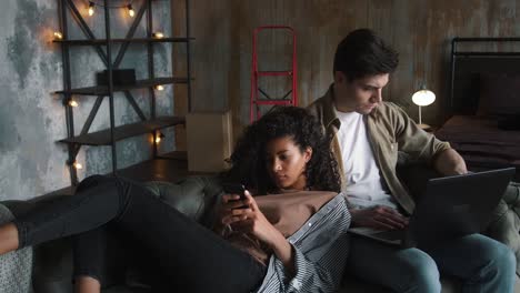 Couple-moving-to-new-apartment.-He-working-at-laptop,-she-using-smartphone.-Relaxing-together-on-gray-couch-in-room-with-cardboard-boxes.-Close-up