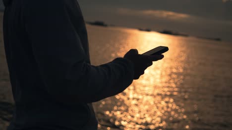 Close-up-athlete-runner-using-smartphone-typing-message-during-resting-after-running-exercising-outdoors-on-the-beach-at-beautiful-sunset-in-summer.