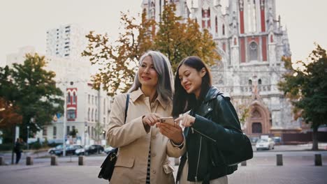 Asiatische-reife-Mutter-und-erwachsene-Tochter-verwenden-Smartphone,-während-sie-auf-dem-Stadtplatz-stehen,-lächelnd-und-auf-etwas-zeigen