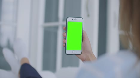 Woman-using-mobile-phone-vertical-green-screen.-Female-holding-in-hand-portable-gadget-close-up-indoors-home-living-room.-Mock-up-for-tracking-or-watching-content.-Blurred-room-on-the-background