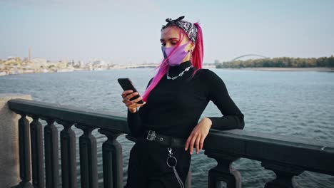 Hipster-woman-in-informal-outfit-and-protective-mask-is-using-her-cellphone-while-leaning-on-fence-of-city-embankment