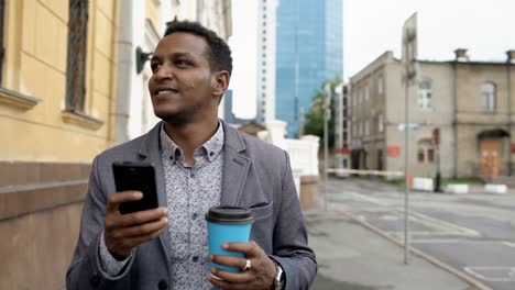 Steadicam-shot-of-young-happy-businessman-using-smartphone-and-walking-with-cup-of-coffee-outdoors